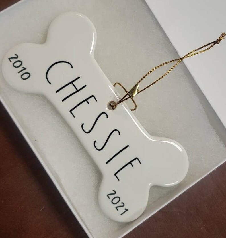 May include: White ceramic bone-shaped ornament with the name "Chessie" and the years "2010" and "2021" printed on it. The ornament has a gold string attached for hanging.