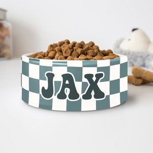 May include: A white and teal checkered dog bowl filled with brown kibble. The bowl has the name "JAX" in a retro font. A jar of treats and a stuffed animal are in the background.