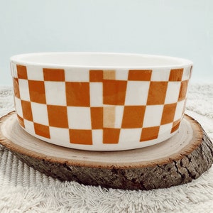 May include: A ceramic bowl with a white and orange checkered pattern. The bowl is cylindrical and sits on a round wooden slice. The background is a light blue color.
