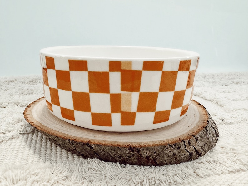 May include: A ceramic bowl with a white and orange checkered pattern. The bowl is cylindrical and sits on a round wooden slice. The background is a light blue color.