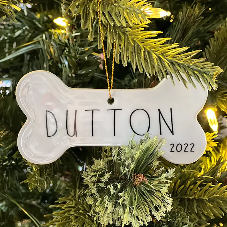 May include: White ceramic bone-shaped Christmas ornament with the name "DUTTON" and the year "2022" written in black.