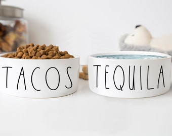 May include: Two white ceramic pet bowls with black lettering. One bowl says "TACOS" and contains brown dog food. The other bowl says "TEQUILA" and contains water.