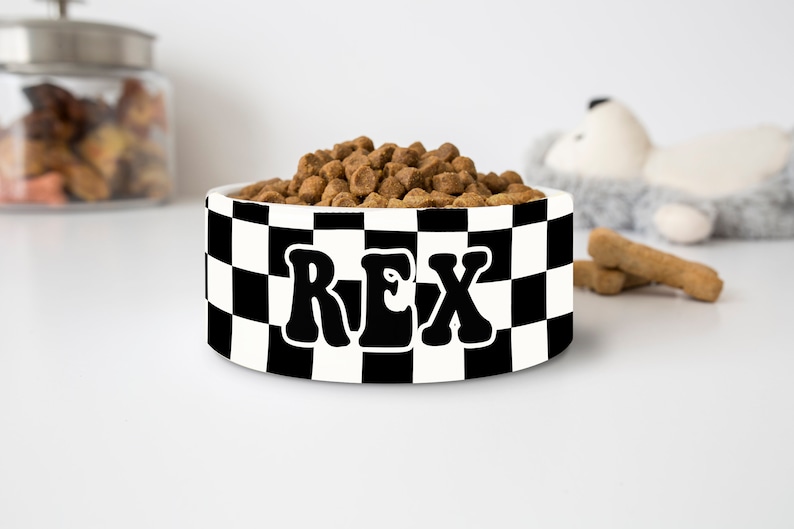 May include: A white ceramic pet bowl with a black and white checkered pattern. The word "REX" is printed in large, black, retro-style letters. The bowl is filled with brown pet food.
