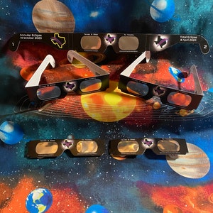May include: Three pairs of eclipse glasses with a black frame and a clear lens. The glasses are labeled "Texas 2 Step" and "To Totality". The glasses are on a blue and purple fabric background with planets and stars.