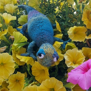 May include: A blue and green knitted chameleon toy with a yellow and green patterned body. The toy is sitting on a bed of yellow flowers.