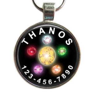 May include: A round, silver-toned Thanos pendant with a black background. The pendant features the word "THANOS" in white, and six colorful gems. A phone number is printed around the edge of the pendant.
