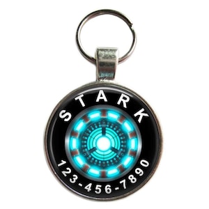 Puede incluir: Un llavero plateado con un dije redondo negro y plateado. El dije tiene un dise&ntilde;o de reactor de arco de Iron Man azul y rojo con la palabra "STARK" y los n&uacute;meros "123-456-7890" alrededor.
