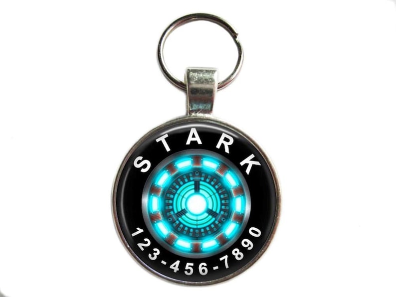 Puede incluir: Un llavero plateado con un dije redondo negro y plateado. El dije tiene un dise&ntilde;o de reactor de arco de Iron Man azul y rojo con la palabra "STARK" y los n&uacute;meros "123-456-7890" alrededor.