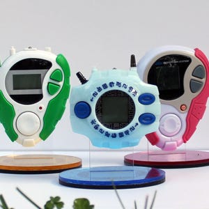 Puede incluir: Tres dispositivos digitales para mascotas de diferentes colores, cada uno con un diseño diferente. Los dispositivos están en soportes de exhibición transparentes. Los dispositivos son blancos, azules y rosados.