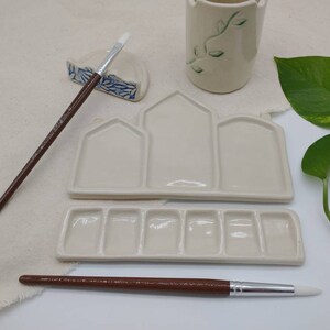 May include: A white ceramic paint palette with three large wells and six smaller wells. The palette is designed to look like a row of houses. Two paintbrushes are included, one with a brown handle and one with a white handle.