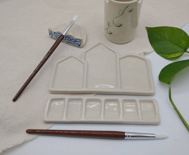 May include: A white ceramic paint palette with three large wells and six smaller wells. The palette is designed to look like a row of houses. Two paintbrushes are included, one with a brown handle and one with a white handle.