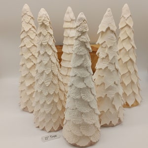 Könnte beinhalten: Sechs weiße Keramik-Weihnachtsbäume, jeder mit einem anderen strukturierten Design, stehen auf einer Holzoberfläche. Die Bäume sind 28 cm hoch und mit "11" Tree" beschriftet.