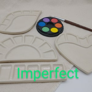 May include: Four ceramic paint palettes in different shapes: a teardrop, a half-circle, a heart, and a rectangular palette with multiple wells. The palettes are white and are arranged on a white fabric surface. A small round palette with eight paint wells is also visible, along with a brown paintbrush.