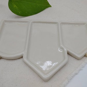 May include: A white ceramic palette with three sections, shaped like a house. The palette is on a white fabric surface.