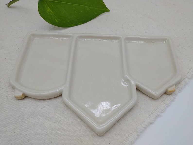 May include: A white ceramic palette with three sections, shaped like a house. The palette is on a white fabric surface.