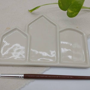 May include: White ceramic paint palette with three wells in the shape of houses. The palette is on a white fabric surface with a green plant in the background. A brown paintbrush is lying on the fabric surface.