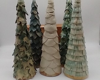 Op de afbeelding: Een verzameling van vijf conische keramische sculpturen in de vorm van bomen. De sculpturen hebben gevarieerde texturen, waaronder gelaagde bladachtige schubben en gegolfde randen, afgewerkt in tinten saliegroen, donker bosgroen en gebroken wit.