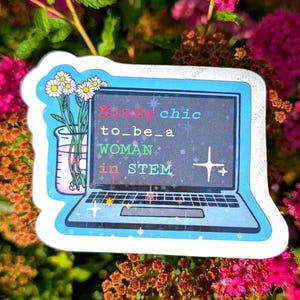 Może przedstawiać: Naklejka przedstawiająca laptopa z ciemnym ekranem, na którym widnieje napis Kinda chic to_be_a WOMAN in STEM kolorowymi literami. Obok laptopa znajduje się szklana zlewka wypełniona różowym płynem oraz bukiet białych stokrotek z żółtymi środkami.