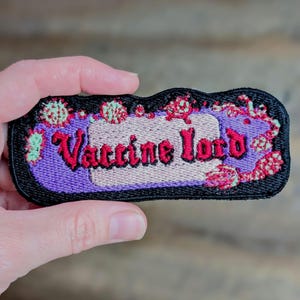 Puede incluir: Un parche bordado rectangular con borde negro, que presenta el texto Vaccine Lord en letras góticas rojas. El fondo incluye un centro morado, un acento rectangular de color beige claro y motivos florales decorativos en rojo y verde.