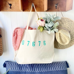 May include: A white canvas tote bag with teal blue numbers 07676 printed on the front. The bag is hanging on a wooden shelf with a pink flower and a straw hat.