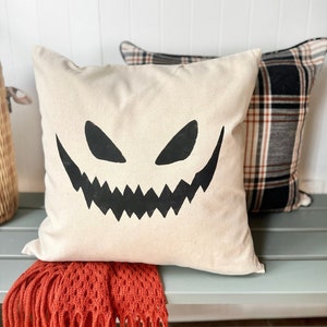 May include: A white pillow with a black jack-o'-lantern face design. The pillow is on a light blue bench with an orange fringed throw blanket.