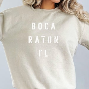 May include: A beige sweatshirt with the words "BOCA RATON FL" printed in white, stencil-style lettering. The sweatshirt has a crew neck and long sleeves. The overall style is casual and comfortable.