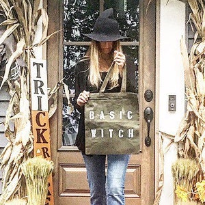 May include: A woman wearing a black witch hat and a black jacket holds a canvas tote bag with the text "BASIC WITCH" printed on it. The bag is hanging from her shoulder. She is standing in front of a door with a "TRICK OR TREAT" sign.