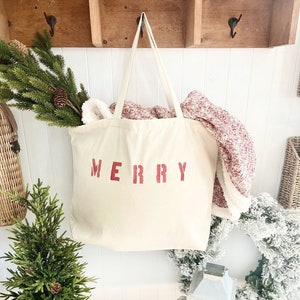May include: A white canvas tote bag with the word "MERRY" printed in red on the front. The bag is hanging on a wooden hook and has a red and white blanket inside.