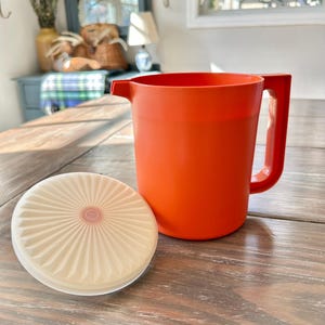 May include: An orange plastic pitcher with a handle and a matching lid. The lid is off to the side, showing a cream-colored, ridged design. The pitcher is sitting on a wooden table, with a glimpse of a window and other decor in the background.