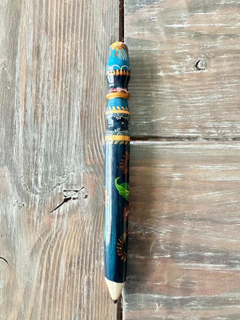 May include: A hand-painted wooden pencil with a white tip. The pencil is dark blue with colorful floral and leaf designs. The top is decorated with blue, orange, and yellow patterns. The pencil is approximately 8 inches long.