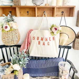 Puede incluir: Una bolsa de lona blanca con las palabras "SUMMER BAG" impresas en rojo. La bolsa est&aacute; colgada de un estante de madera con ganchos. Hay otros art&iacute;culos decorativos en el estante, incluyendo una concha, una figurita de p&aacute;jaro y una cesta de mimbre.
