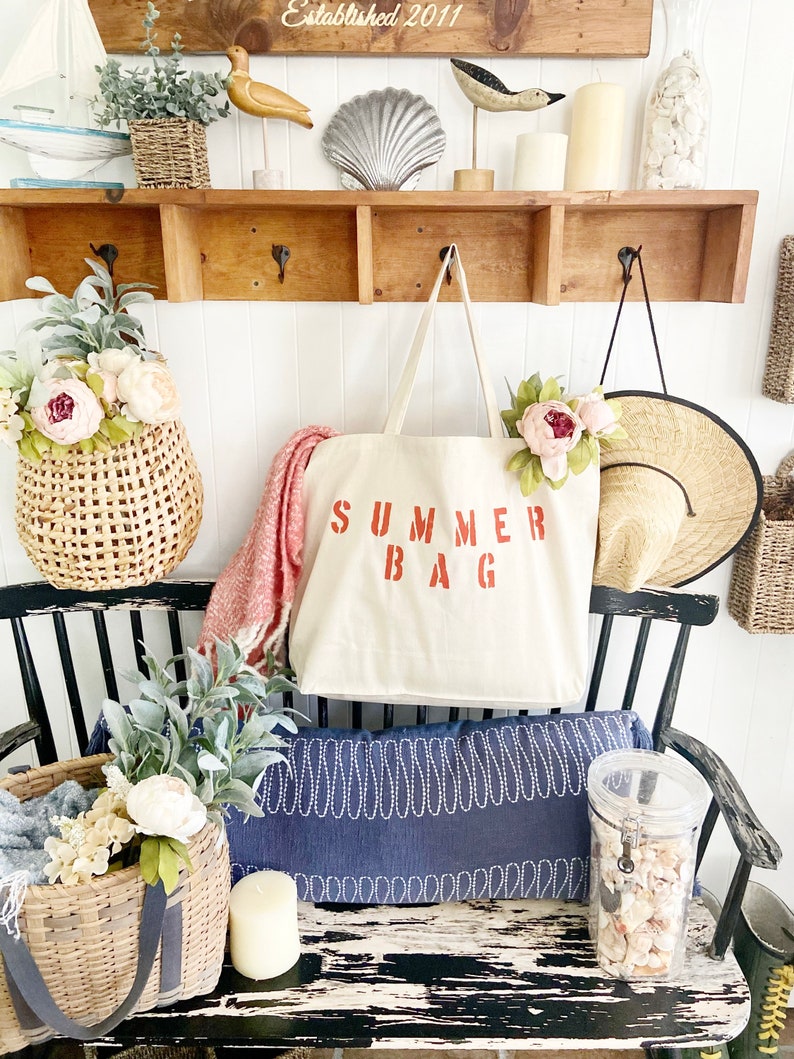 Puede incluir: Una bolsa de lona blanca con las palabras "SUMMER BAG" impresas en rojo. La bolsa est&aacute; colgada de un estante de madera con ganchos. Hay otros art&iacute;culos decorativos en el estante, incluyendo una concha, una figurita de p&aacute;jaro y una cesta de mimbre.