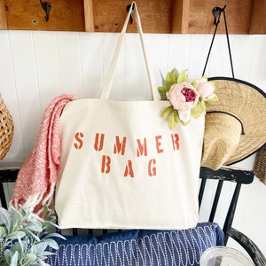 Puede incluir: Una bolsa de lona blanca con las palabras "SUMMER BAG" impresas en naranja. La bolsa est&aacute; colgada de una estanter&iacute;a de madera con una flor rosa y un sombrero de paja.