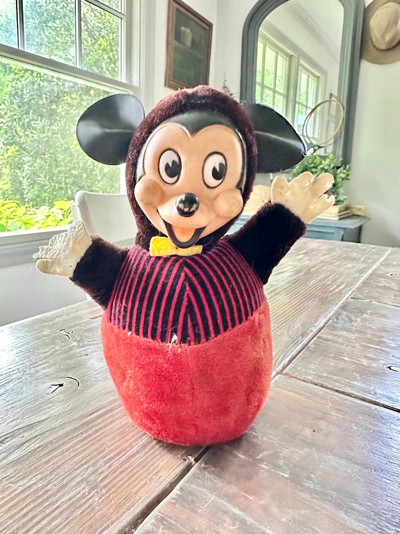 May include: Vintage Mickey Mouse plush toy with a red and black striped torso, red base, and black ears. The toy has a brown hood, a yellow bow tie, and outstretched arms. The toy is sitting on a wooden table.