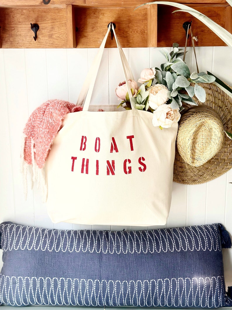May include: A white canvas tote bag with red lettering that says "BOAT THINGS". The bag is hanging on a wooden shelf with a pink and green floral arrangement and a straw hat.