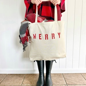 May include: A white canvas tote bag with the word "MERRY" printed in red. The bag is filled with a red and black plaid blanket and is being held by a person wearing a red and black plaid poncho.