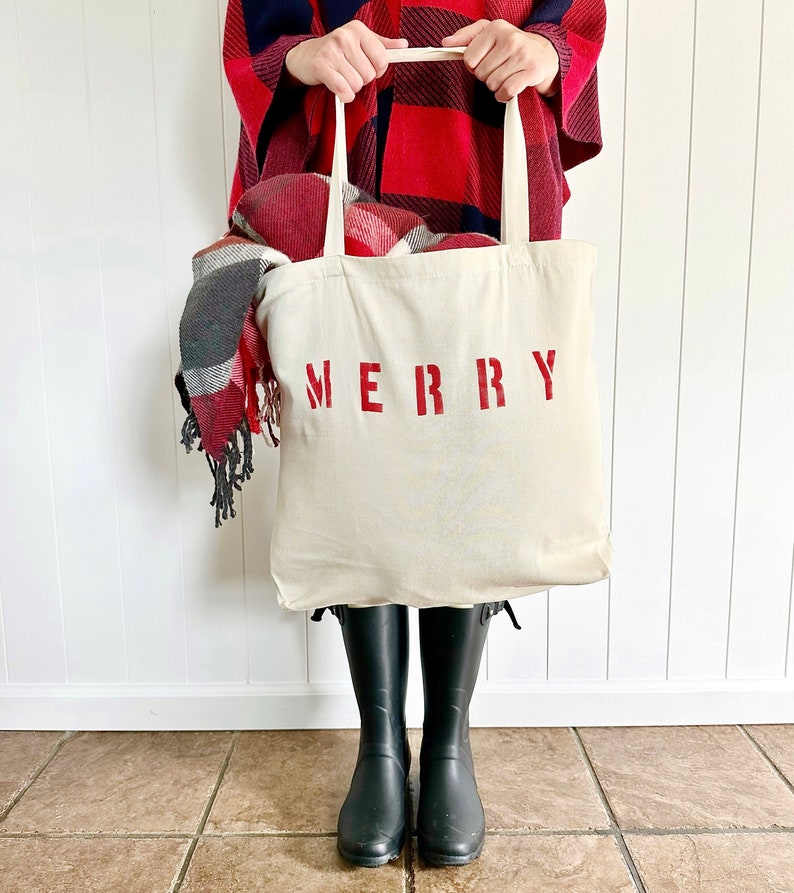 May include: A white canvas tote bag with the word "MERRY" printed in red. The bag is filled with a red and black plaid blanket and is being held by a person wearing a red and black plaid poncho.