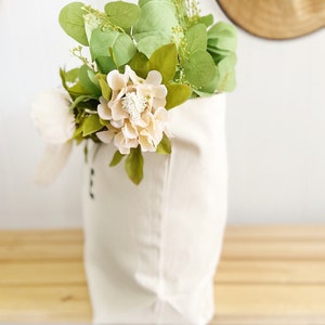 May include: A white canvas tote bag filled with a bouquet of pale pink flowers and green eucalyptus leaves.