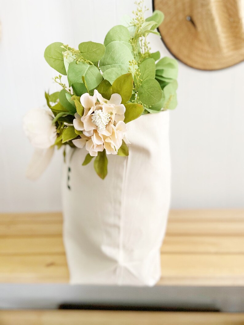 May include: A white canvas tote bag filled with a bouquet of pale pink flowers and green eucalyptus leaves.