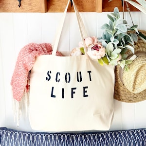 May include: A white canvas tote bag with black text that reads "SCOUT LIFE". The bag is hanging on a wooden shelf with a pink and white blanket and a green plant in the background.