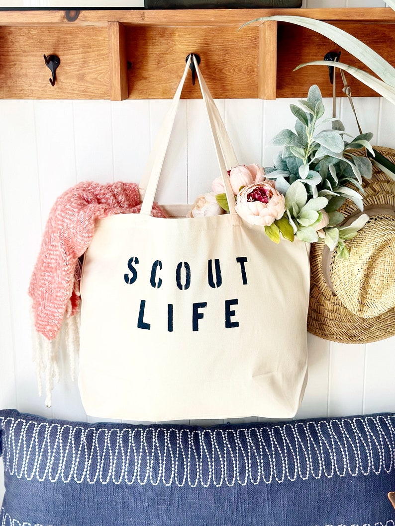 May include: A white canvas tote bag with black text that reads "SCOUT LIFE". The bag is hanging on a wooden shelf with a pink and white blanket and a green plant in the background.