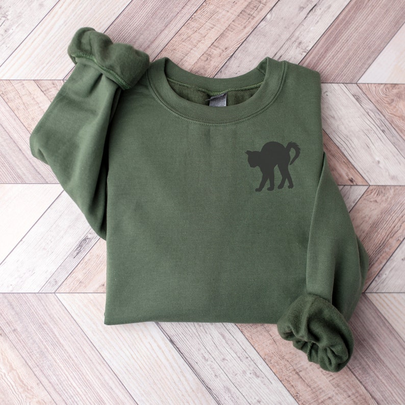 Op de afbeelding: Een groen sweatshirt met een zwart katten silhouet op de voorkant. Het sweatshirt is gevouwen en ligt op een houten oppervlak.