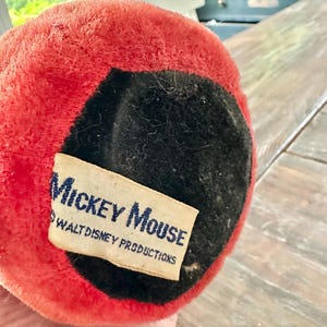 May include: Close-up of a vintage Mickey Mouse plush toy. The toy has a red plush exterior with a black felt base. A rectangular tag with the words "Mickey Mouse" and "Walt Disney Productions" is attached.