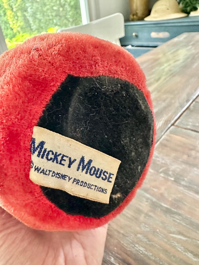May include: Close-up of a vintage Mickey Mouse plush toy. The toy has a red plush exterior with a black felt base. A rectangular tag with the words "Mickey Mouse" and "Walt Disney Productions" is attached.
