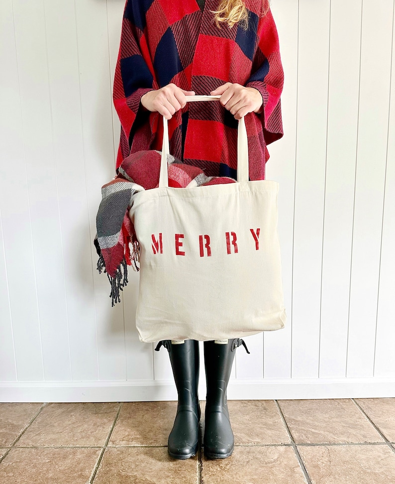 May include: A white canvas tote bag with red lettering that says "MERRY". The bag is being held by a person wearing a red and black plaid poncho and black rain boots.