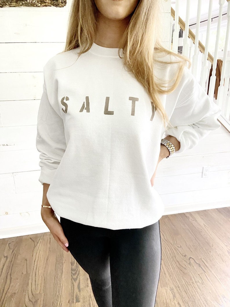 May include: White sweatshirt with the word "SALT" printed in brown on the front. The sweatshirt is being worn by a person with long blonde hair.