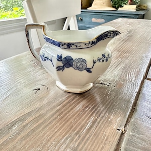 May include: A white ceramic creamer with blue floral patterns and gold trim. The creamer has a handle and a spout, and is sitting on a wooden table. The creamer is a vintage or antique item.