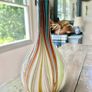 K&ouml;nnte beinhalten: Eine dekorative Glasvase in Tropfenform. Die Vase hat vertikale Streifen in Rot-, Orange-, T&uuml;rkis-, Gr&uuml;n- und Wei&szlig;t&ouml;nen. Der obere Teil der Vase hat ein t&uuml;rkisfarbenes Band mit goldenem Text. Die Vase steht auf einer Holzoberfl&auml;che.