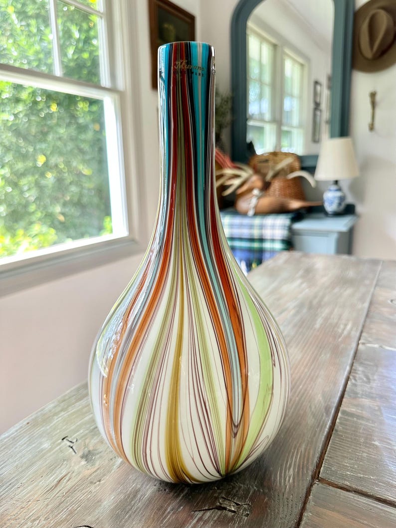 K&ouml;nnte beinhalten: Eine dekorative Glasvase in Tropfenform. Die Vase hat vertikale Streifen in Rot-, Orange-, T&uuml;rkis-, Gr&uuml;n- und Wei&szlig;t&ouml;nen. Der obere Teil der Vase hat ein t&uuml;rkisfarbenes Band mit goldenem Text. Die Vase steht auf einer Holzoberfl&auml;che.