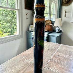 May include: A tall, decorative wooden pencil with a painted design. The pencil is primarily dark blue with colorful floral and geometric patterns. The tip is a light beige color, and the pencil stands upright on a wooden table.